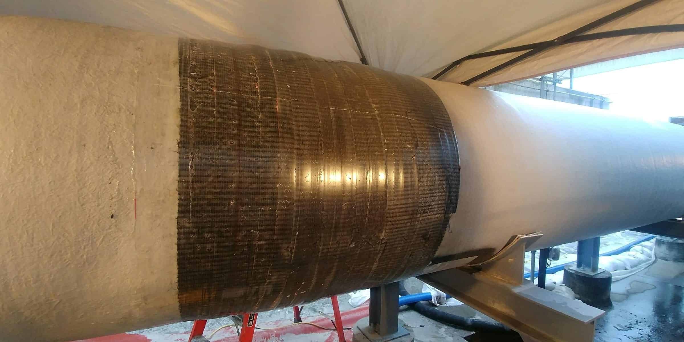 Large diameter pipe inside a white containment tent, part-coated in beige and wrapped with a wide dark composite band
