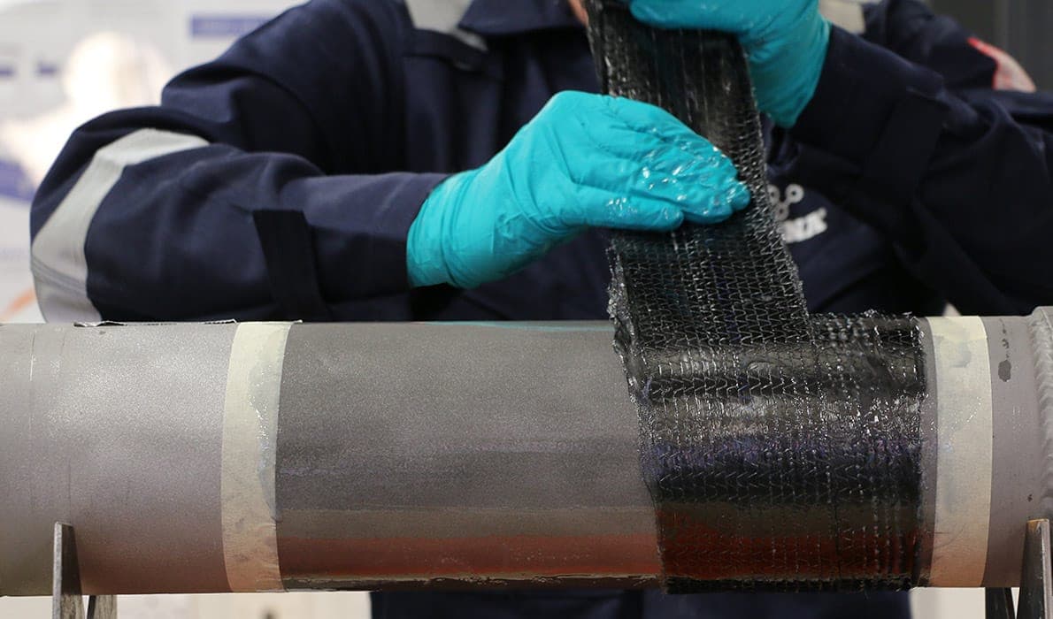 Gloved hands laying a strip of black composite wrap tape onto a gray pipe on a stand
