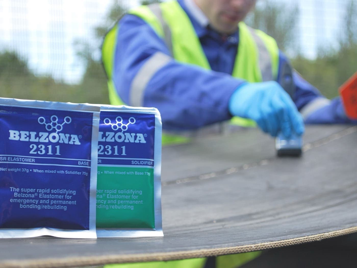 Elastomer sachets in sharp foreground on a conveyor belt, with a technician in blue coveralls and hi-vis working the splice behind