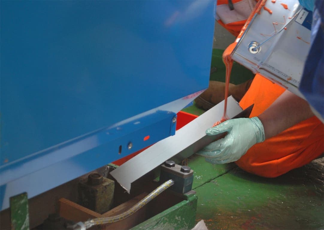 Applicator in orange hi-vis pouring resin from a tin down a machined steel shim beneath a blue machine base