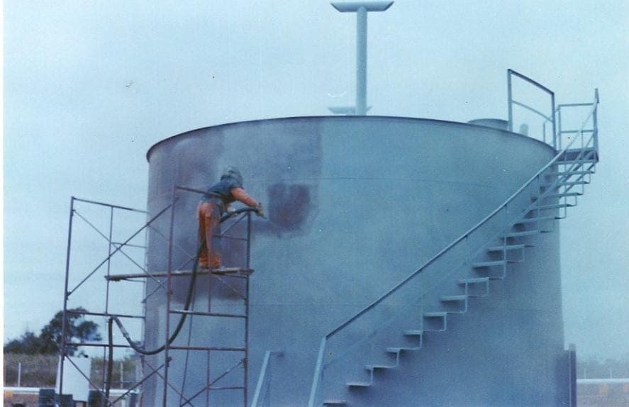 Worker on light scaffold spray-applying coating to the shell of a large outdoor storage tank with an external spiral stair