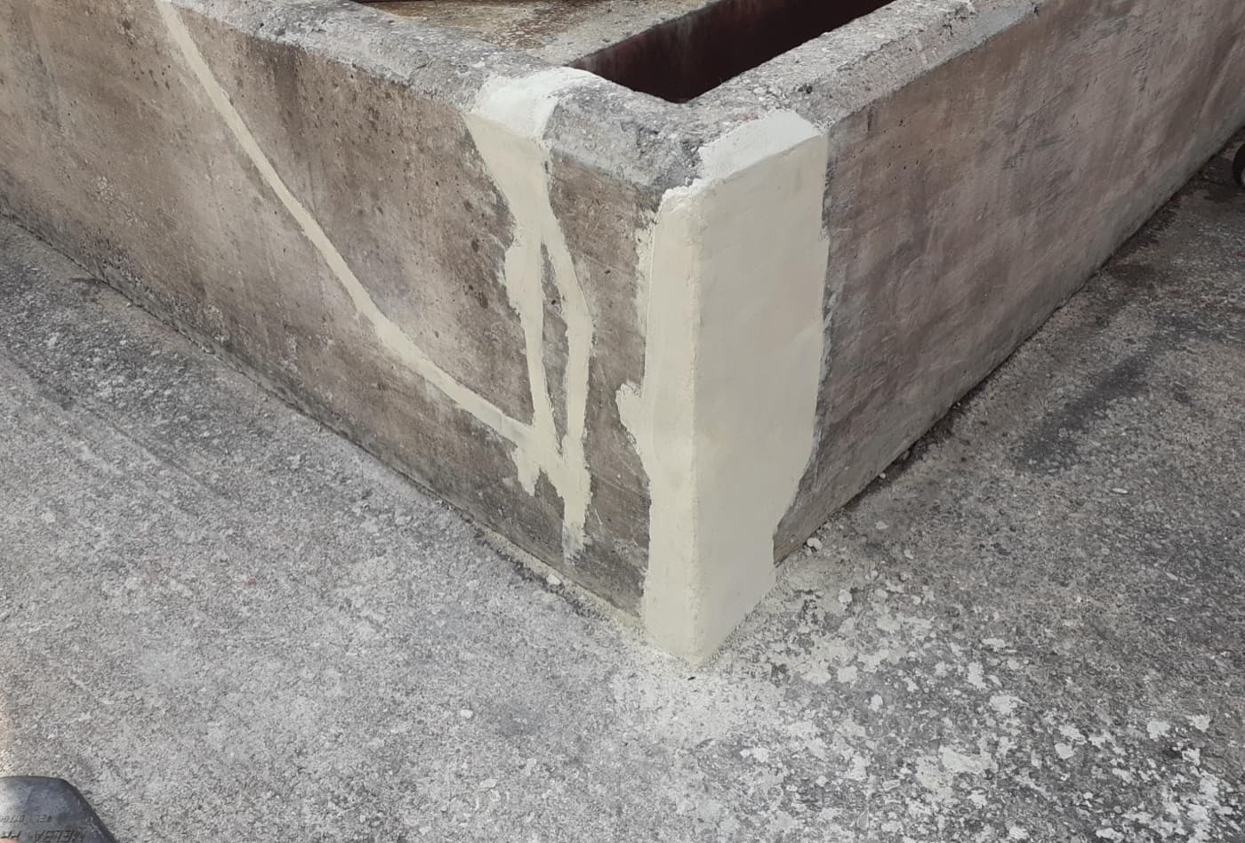 Low concrete bund wall corner with cracks cut out and filled with pale mortar, the corner arris rebuilt