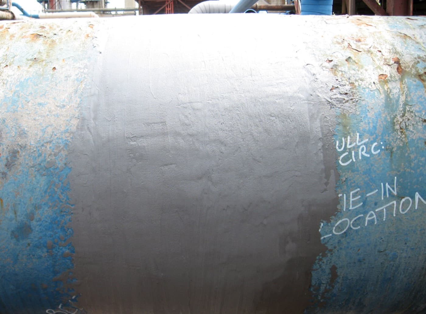 Large horizontal pipe with flaking blue paint and surface corrosion, a wide gray composite wrap applied around the center; chalk notes read "full circ" and "tie-in location"