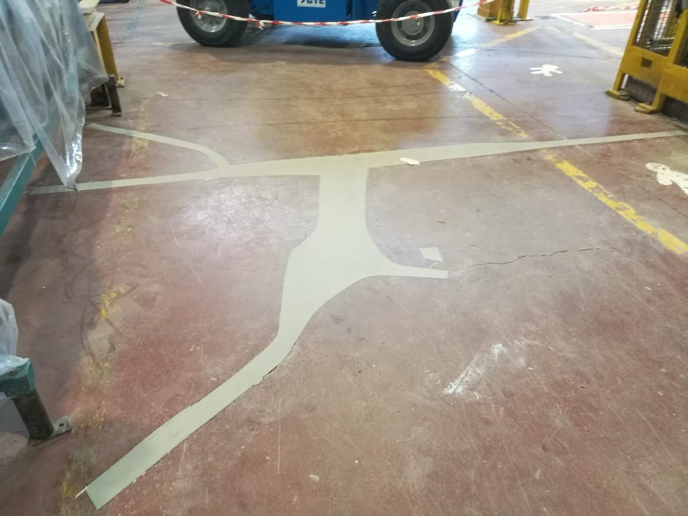 Factory floor coated red-oxide with a long pale repair band filling a crack across the slab; a lift truck and barrier tape behind