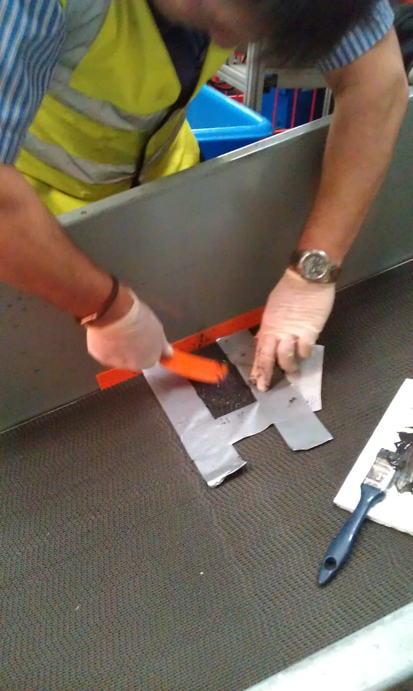 Worker in high-visibility clothing and gloves brushing black repair compound into a taped patch on a conveyor belt in place
