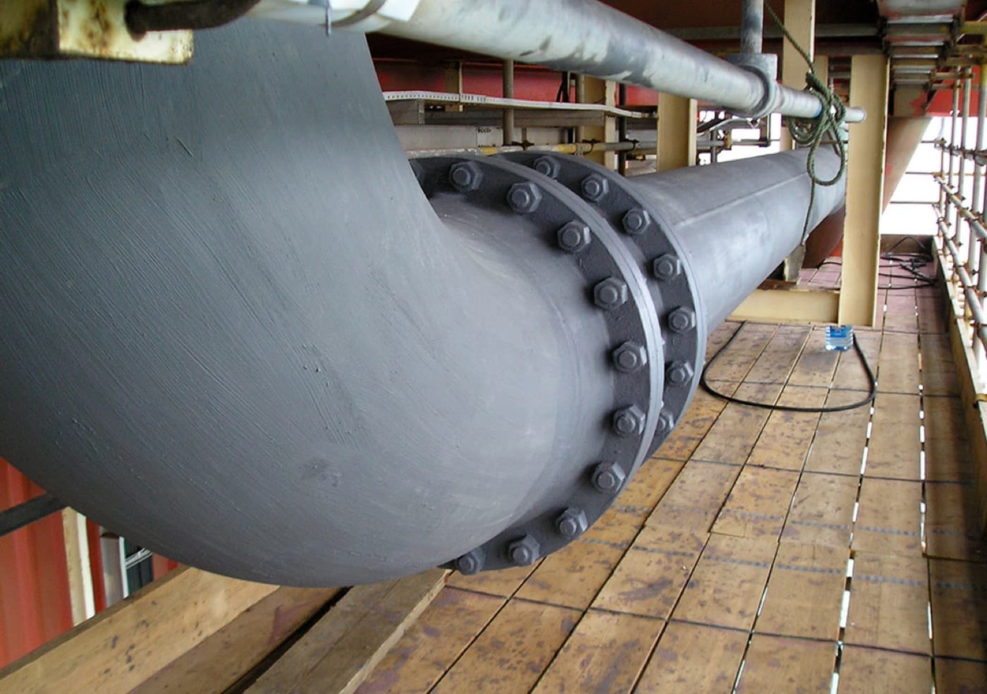 Large process pipework coated uniform gray, including a bolted flange joint, seen from a scaffold boarded walkway
