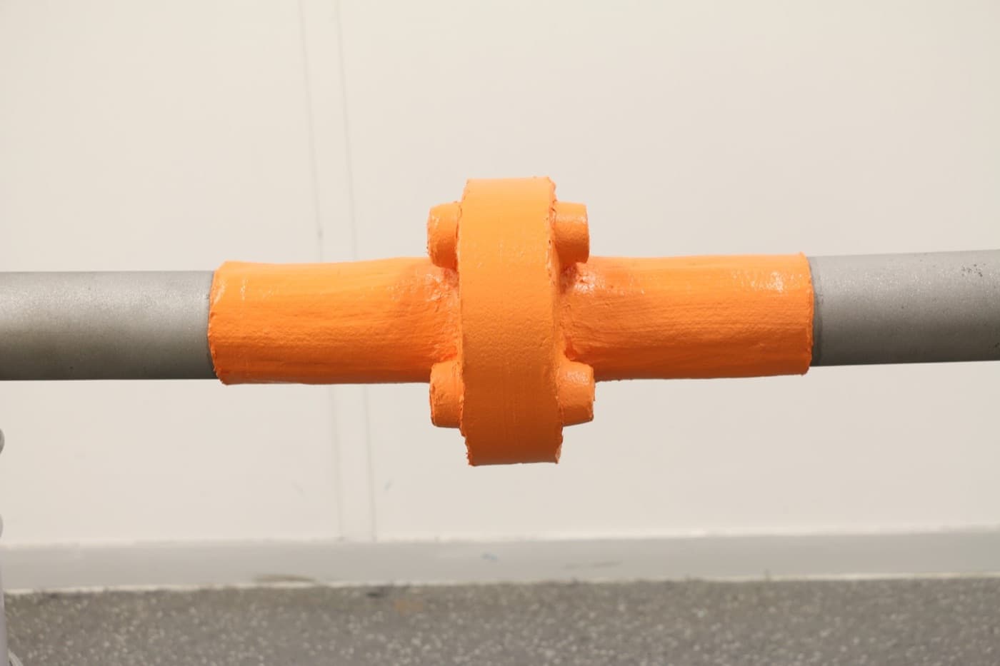Gray pipe with a bolted flanged coupling, the center section coated bright orange against a white wall