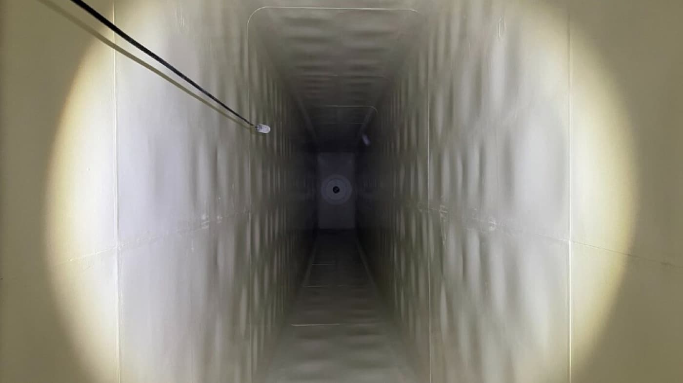 Interior of a long rectangular tank chamber lined in pale cream, lit by two work lamps