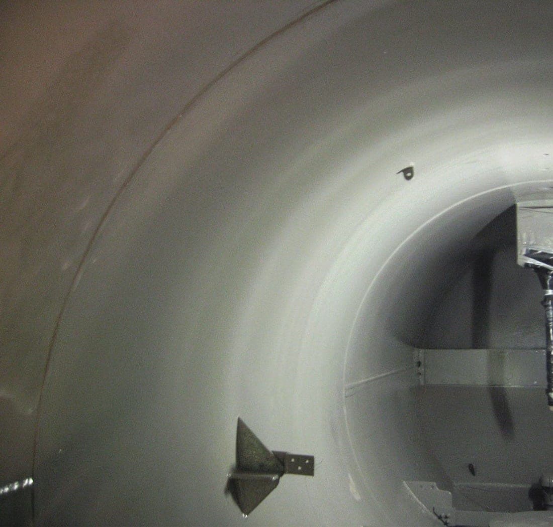 Interior of a large cylindrical vessel freshly coated pale gray, with a worker in red coveralls and a dust mask at the right-hand edge