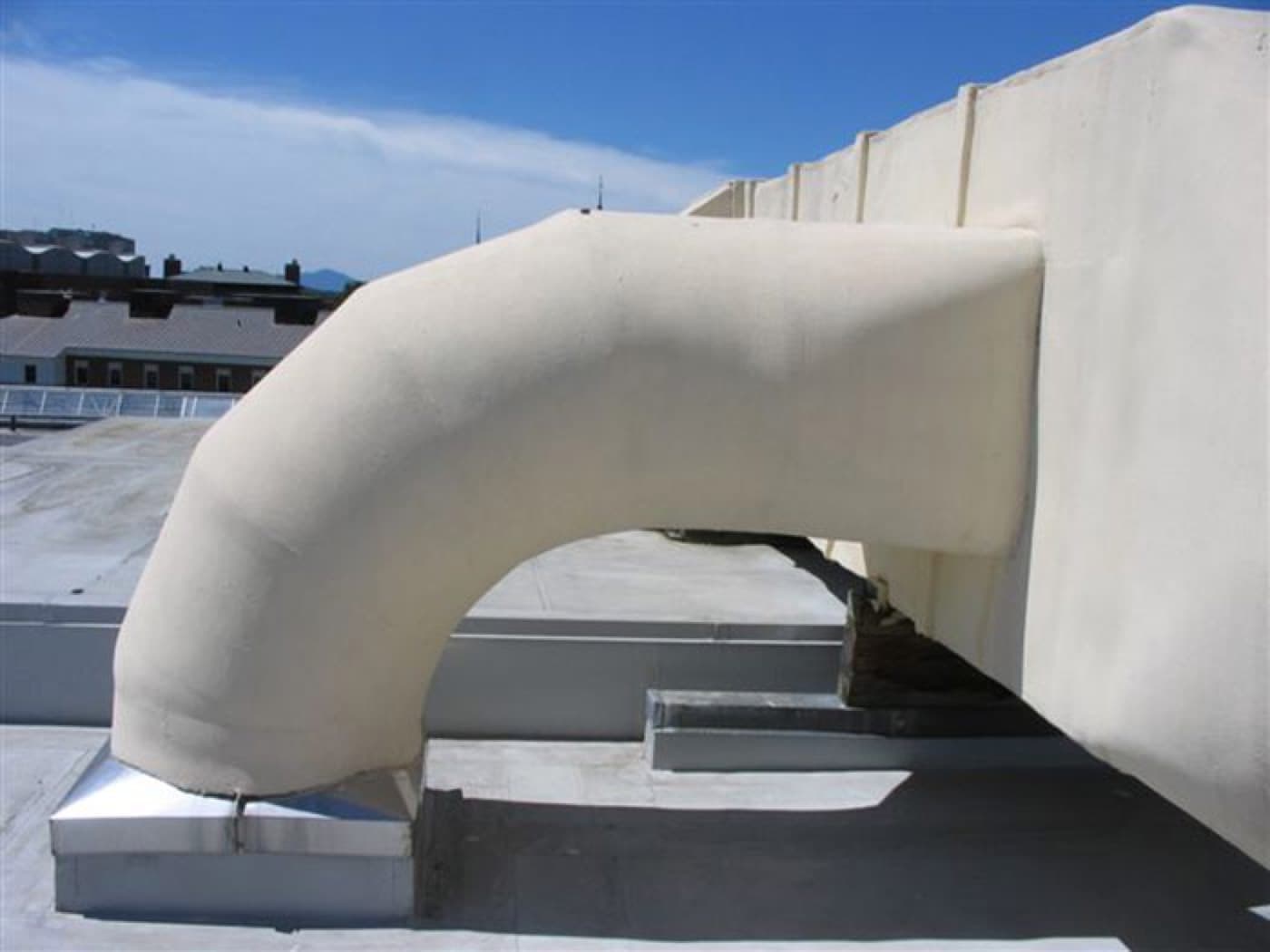 Large ductwork elbow on a rooftop, coated cream-white, against a clear blue sky