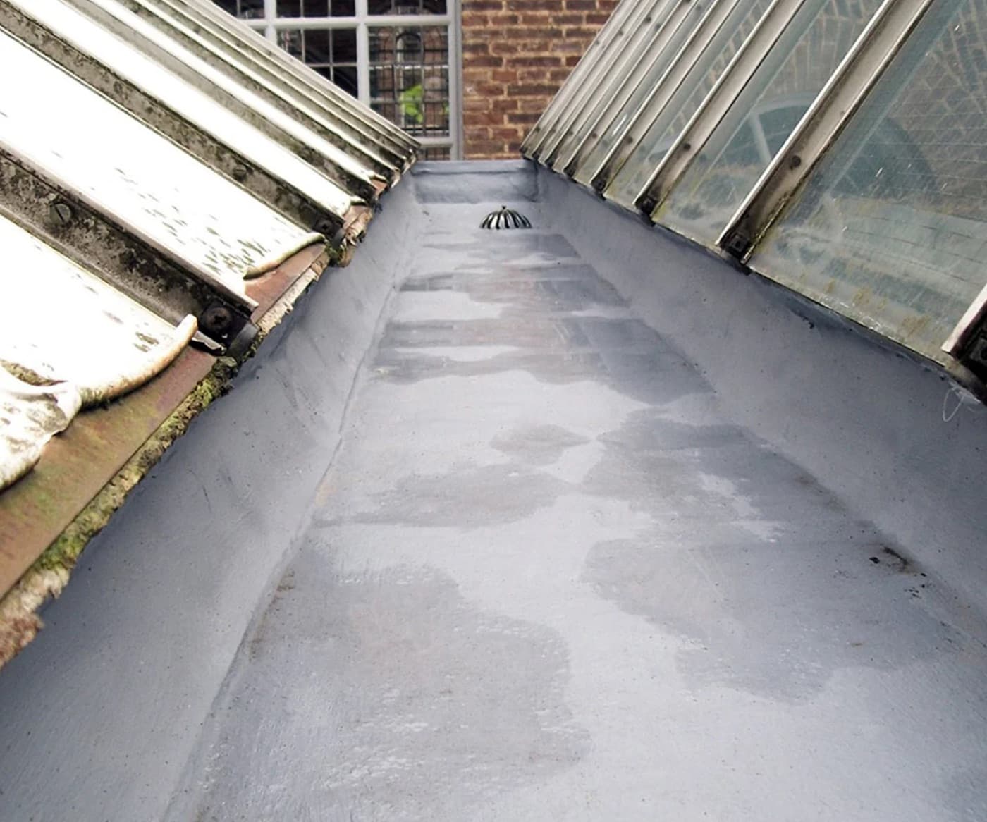 Gray liquid-applied membrane in a roof valley between two glazed rooflights, with a domed rainwater outlet
