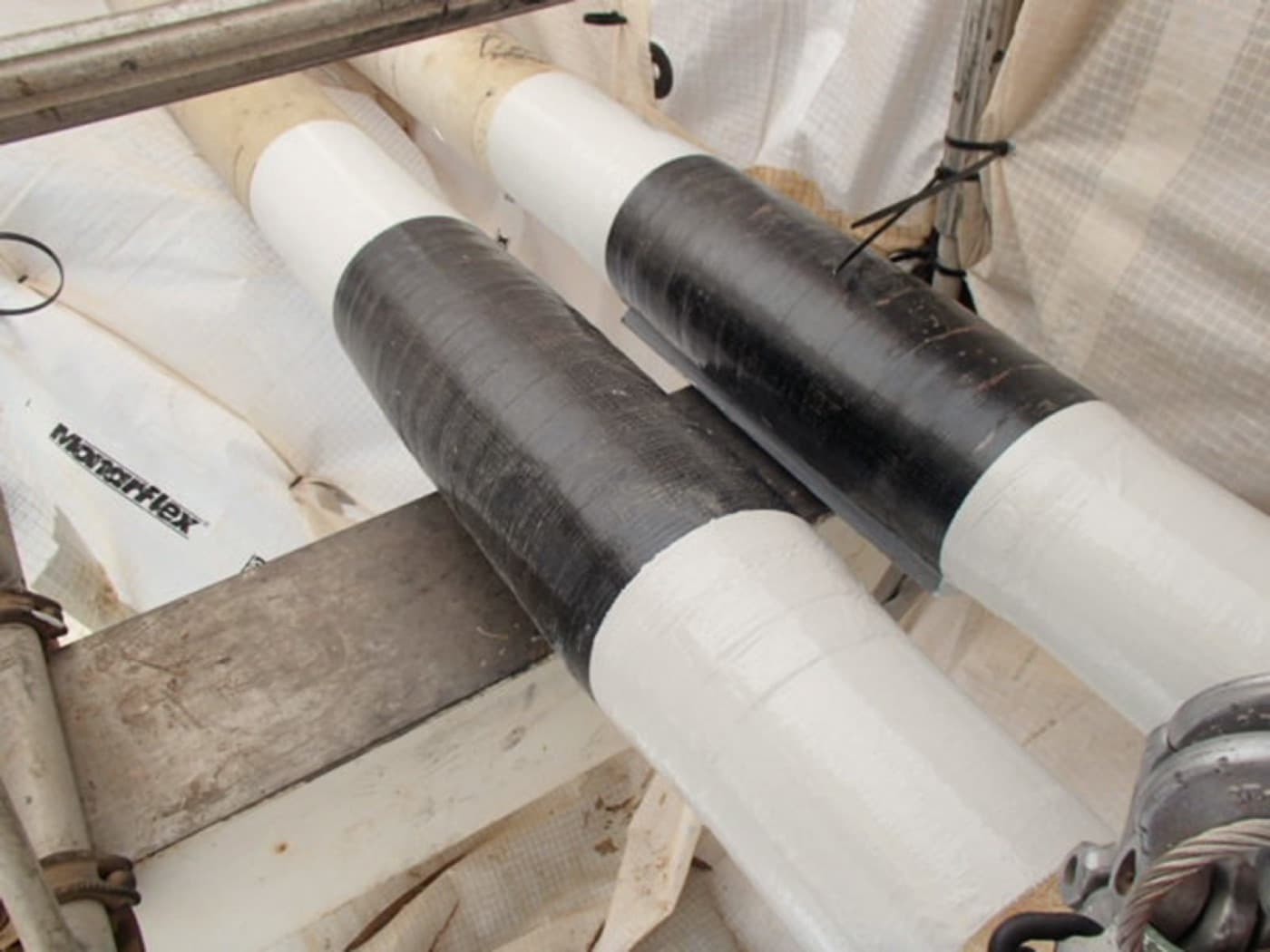 White-lagged pipework on scaffold with two black composite wrap bands applied over the line, under white containment sheeting