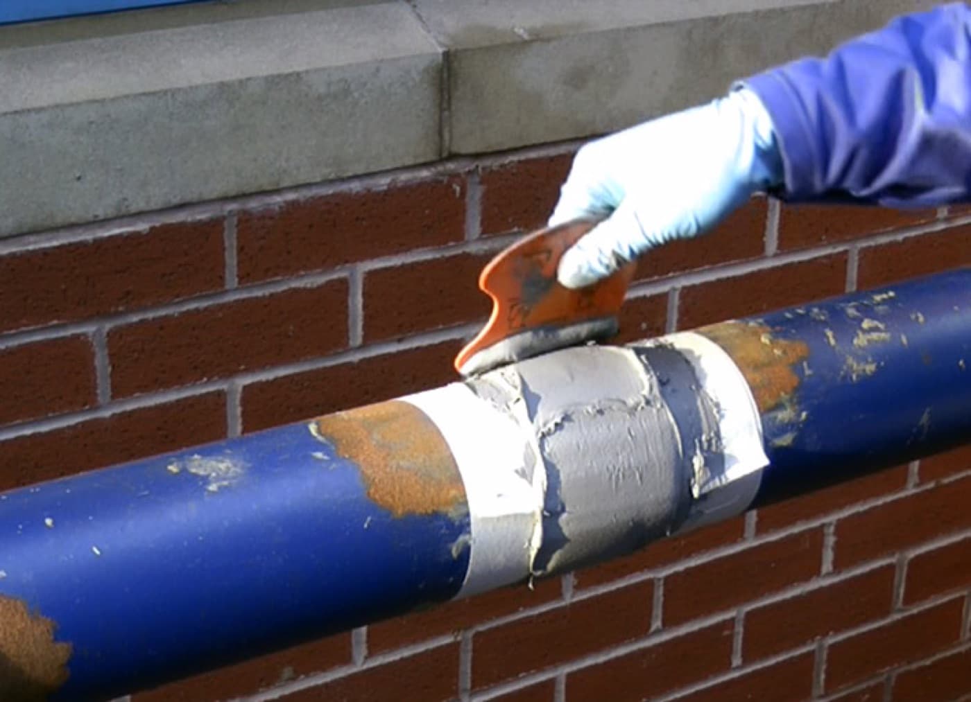 Blue painted pipe on a brick wall with corroded patches; a masked section is being filled with gray paste using an orange spreader