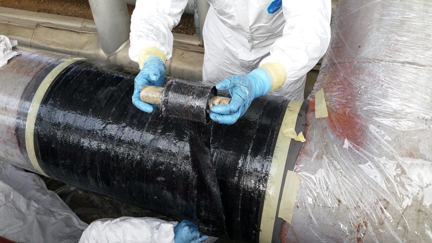 Two workers in disposable coveralls and gloves applying black composite wrap tape to a large insulated pipe inside a containment enclosure
