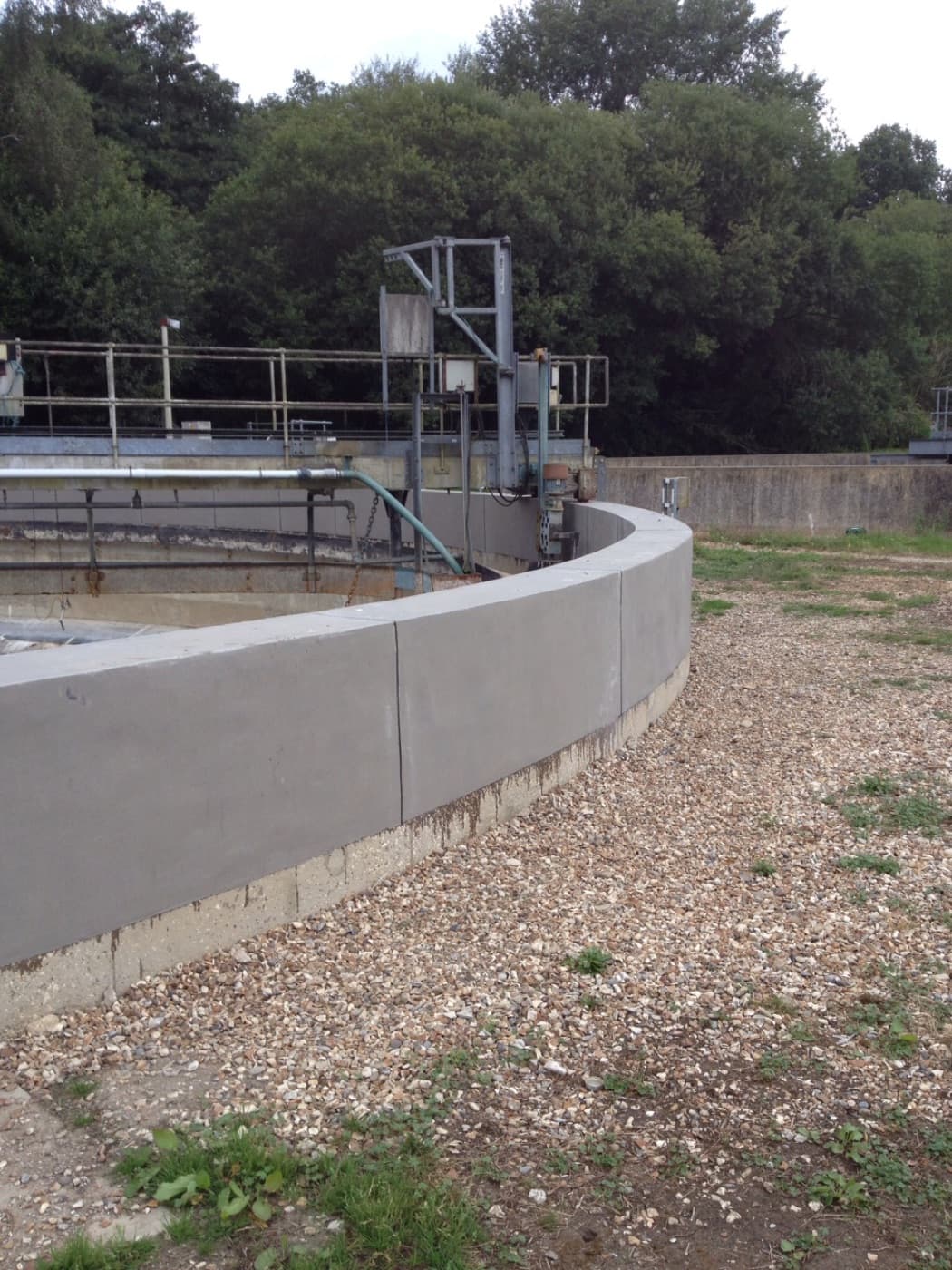 Circular clarifier at a water treatment works, its curved channel wall resurfaced in smooth gray, with gravel and trees beyond