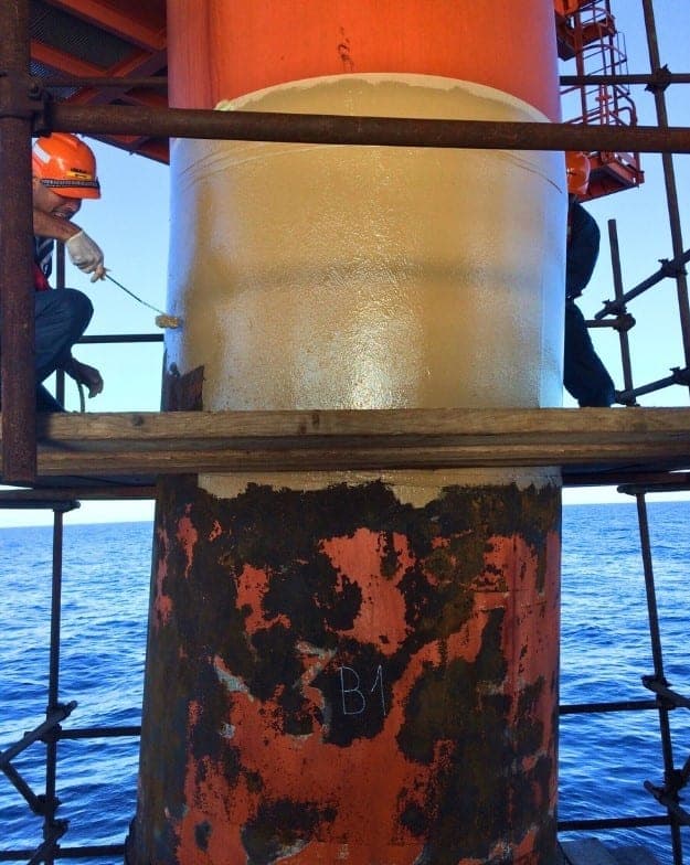 Offshore platform leg above open sea, the lower half heavily rusted and marked "B1", the upper half freshly coated pale gold, with two workers on scaffold