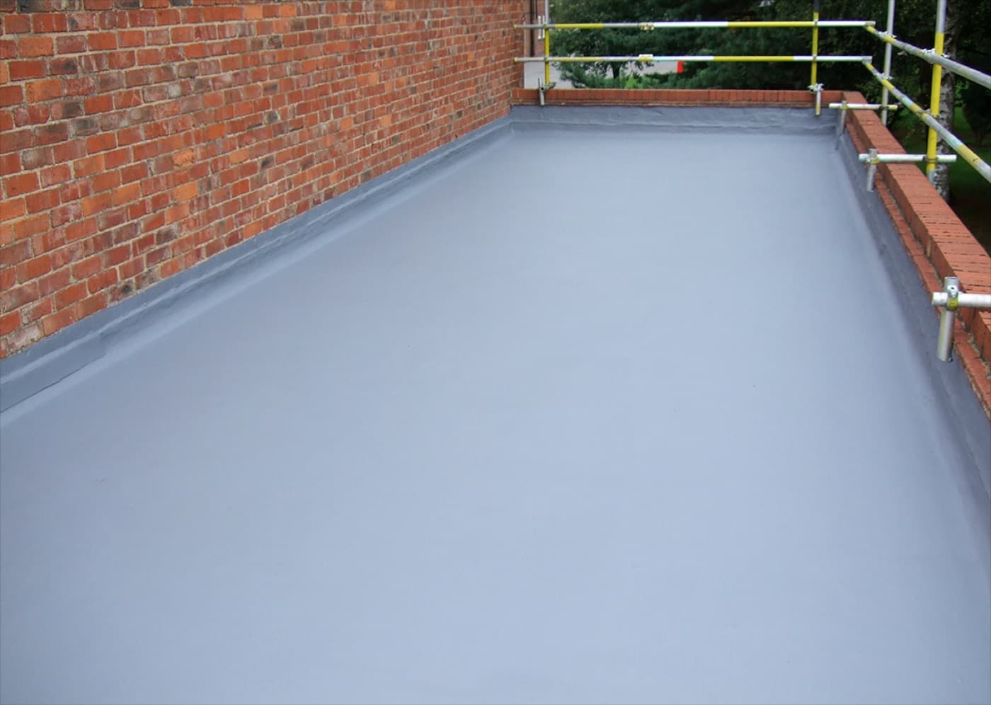 Flat roof finished in gray liquid coating, bounded by a red brick parapet with scaffold standards beyond
