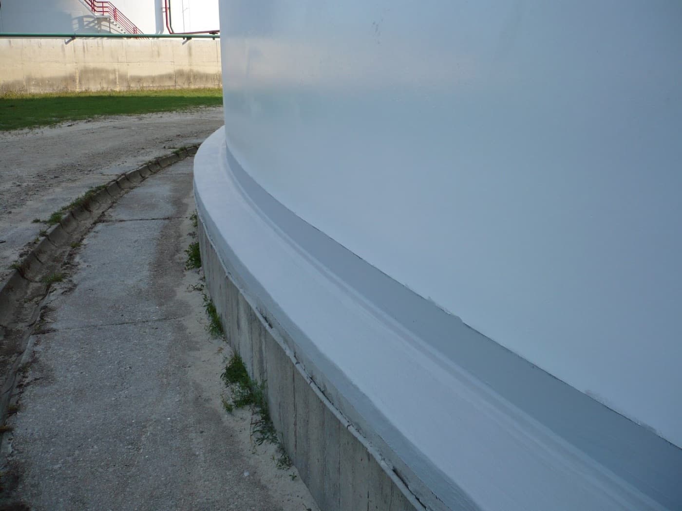 Base of a white storage tank with a gray sealing fillet formed around the chime onto the concrete ringwall, tank farm beyond
