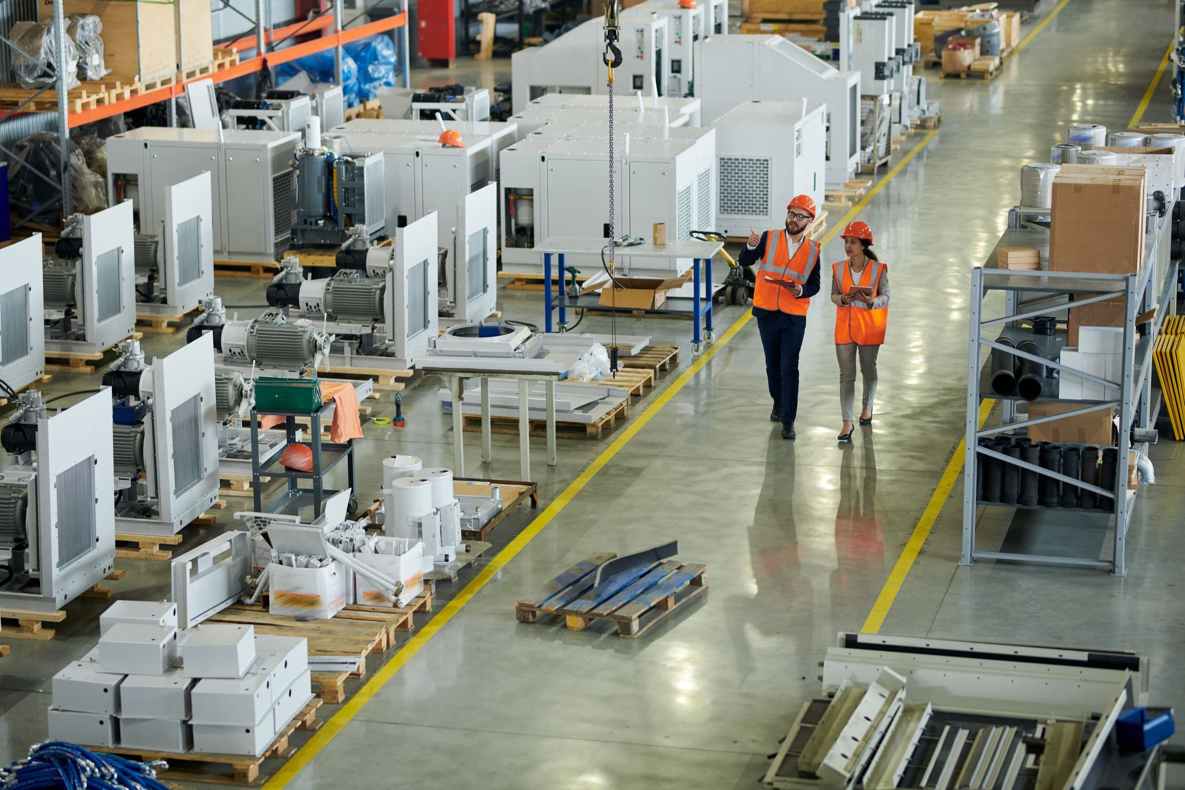 Two people in orange hi-vis vests and hard hats walking a plant aisle between pallets of equipment