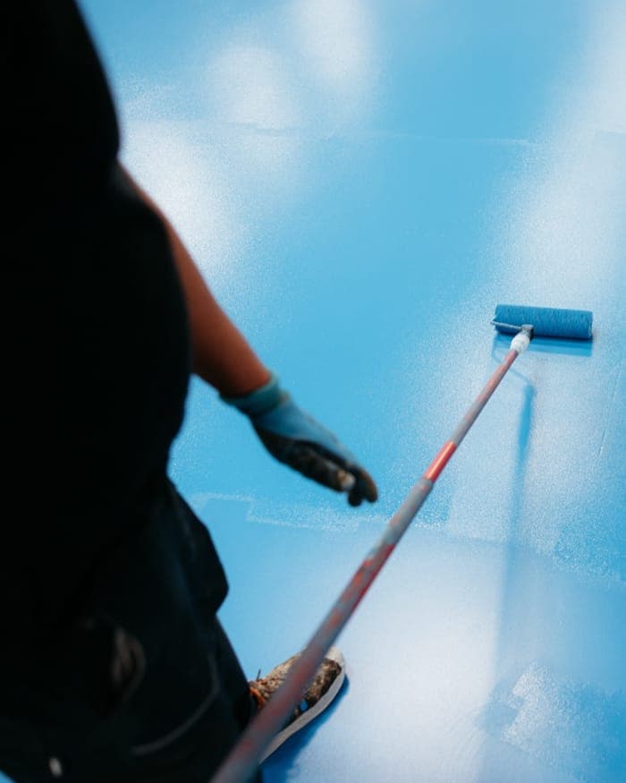 Person seen from behind rolling a bright blue coating across a large floor with a long-handled roller