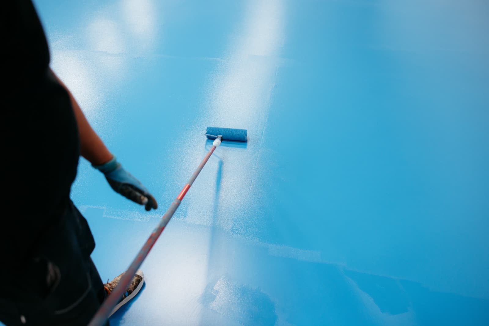 Person seen from behind rolling a bright blue coating across a large floor with a long-handled roller
