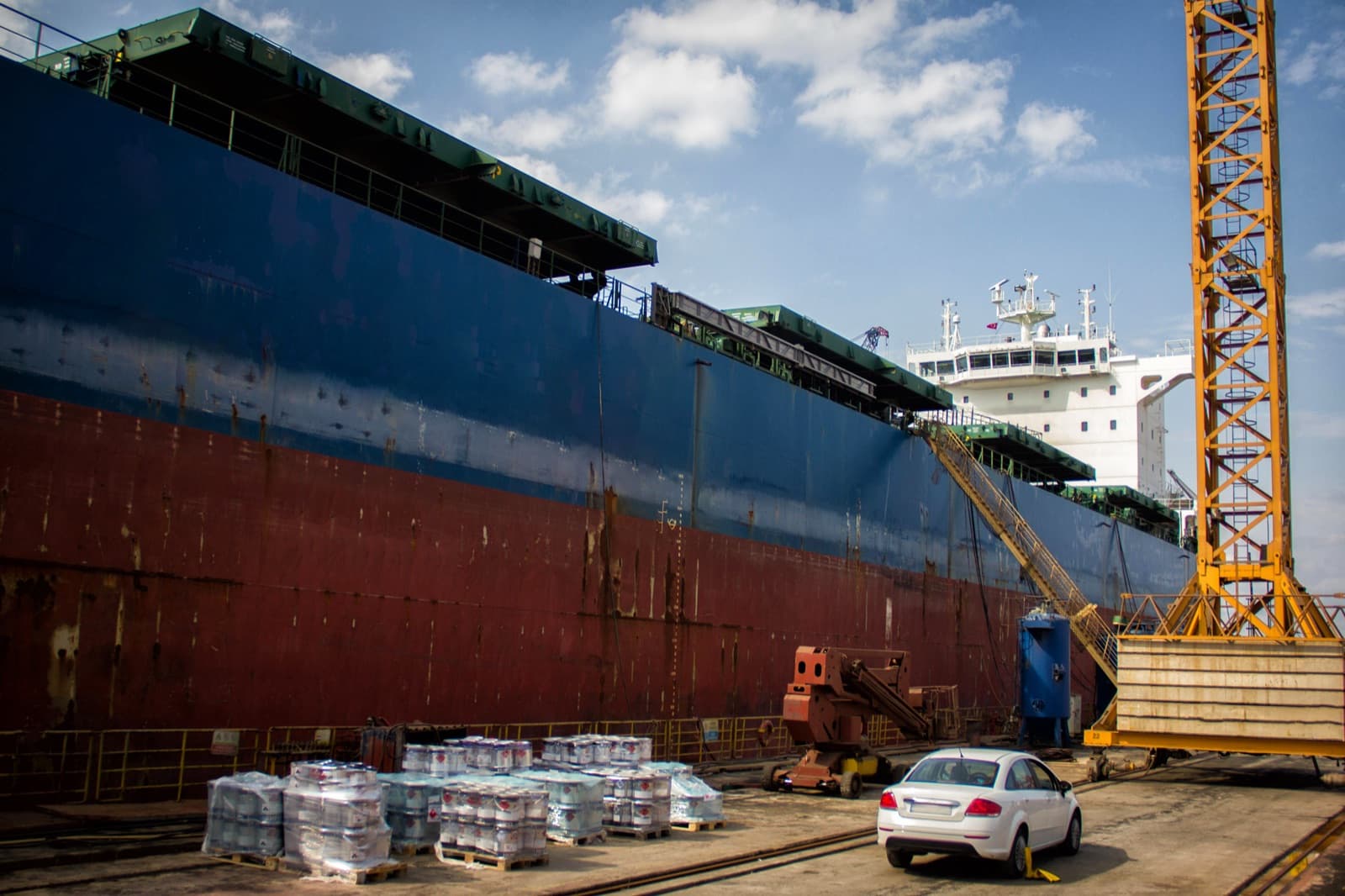 Bulk carrier in dry dock beside a yellow gantry crane, its hull showing blue topsides and red anti-fouling, with pallets of pails on the dock floor